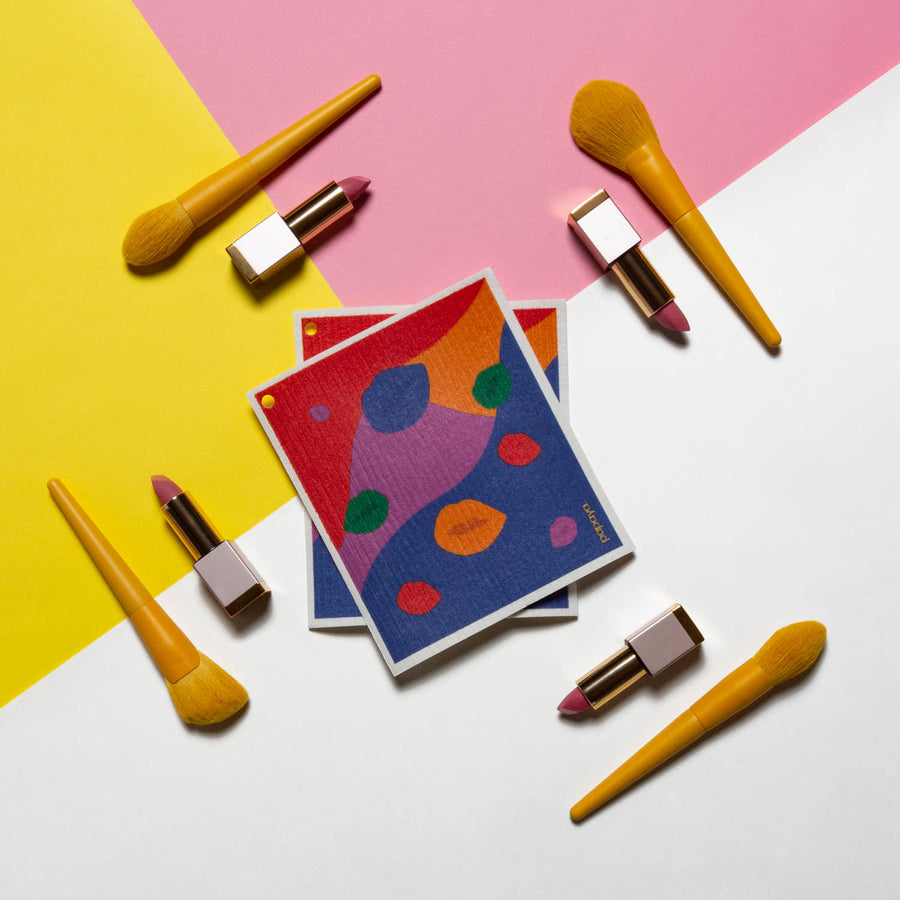 Two reusable paper towels in a colorful lip design surrounded by yellow makeup brushes and lipsticks