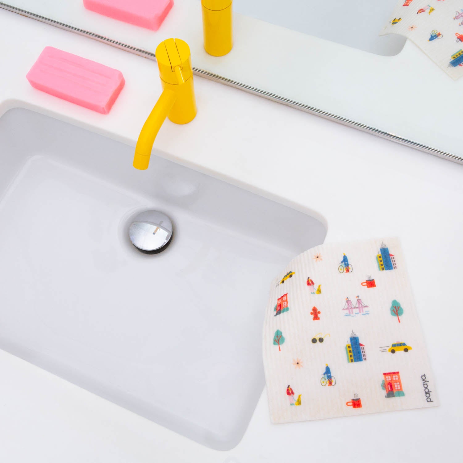 Reusable paper towel with city pattern design hanging over a white sink with a yellow faucet and pink bar of soap