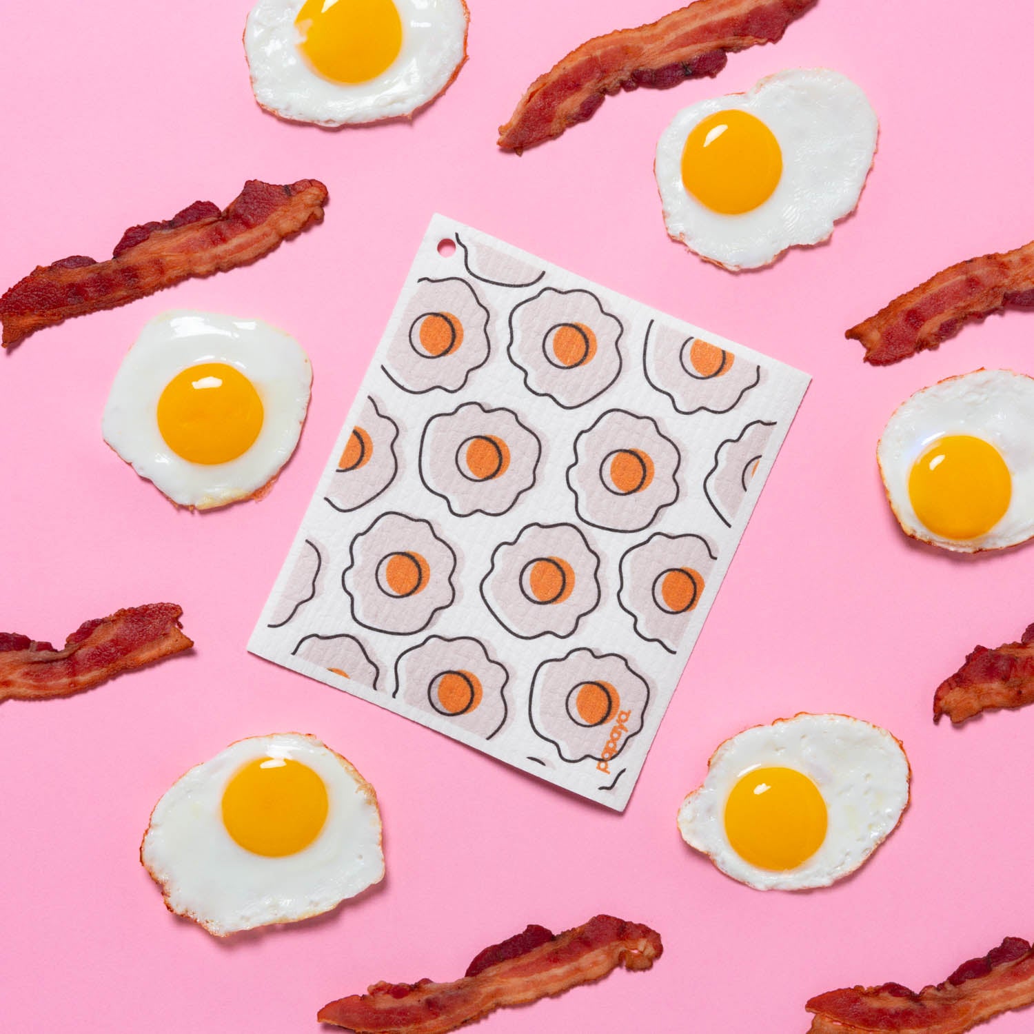 Reusable paper towel with egg design on pink background surrounded by a pattern of sunny side up eggs and bacon